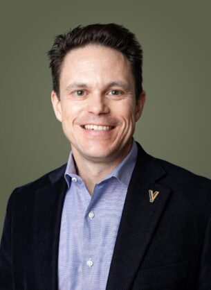 Smiling man in a dark blazer over a light blue shirt, olive background, close-up portrait.