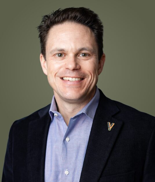 Smiling man in a dark blazer over a light blue shirt, olive background, close-up portrait.