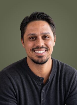 Close-up portrait of a smiling man with dark hair and a black henley, olive green backdrop.