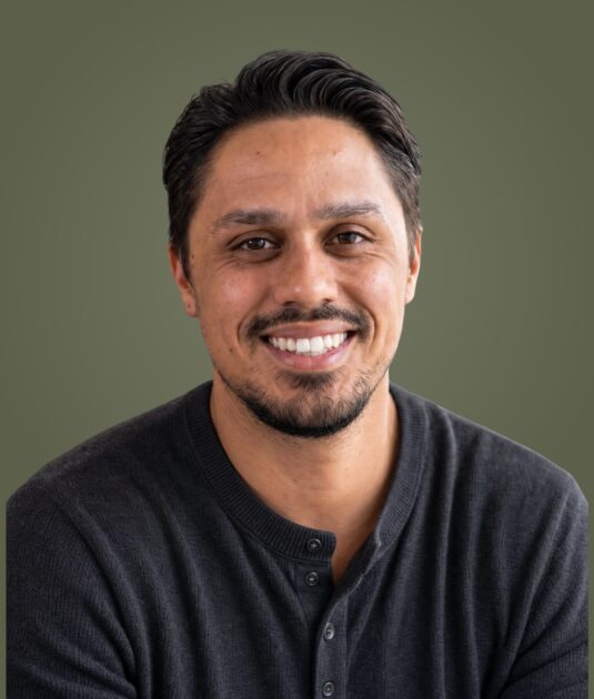 Close-up portrait of a smiling man with dark hair and a black henley, olive green backdrop.