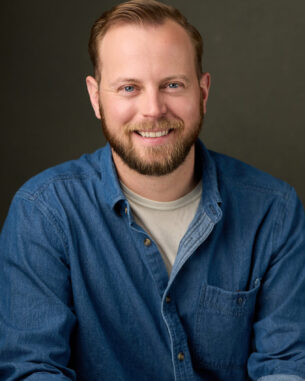 Smiling man in a blue denim shirt posed against a dark background.