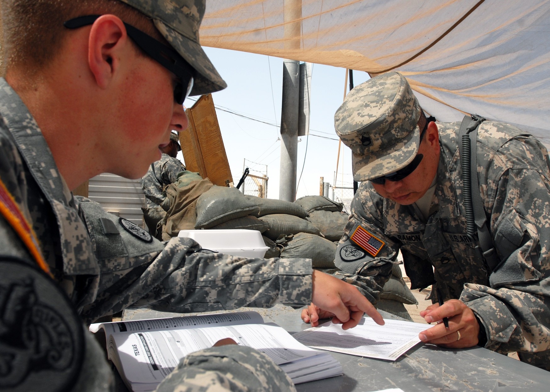 Two soldiers in desert camouflage uniforms review documents on a table under a tarp at a sandbag barrier in a outdoor field setting.