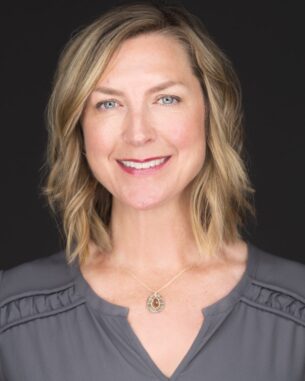 Professional headshot of a smiling blonde woman in a gray blouse, wearing a teardrop pendant necklace, against a dark backdrop.