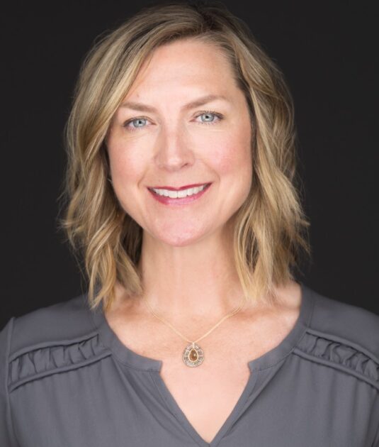 Professional headshot of a smiling blonde woman in a gray blouse, wearing a teardrop pendant necklace, against a dark backdrop.