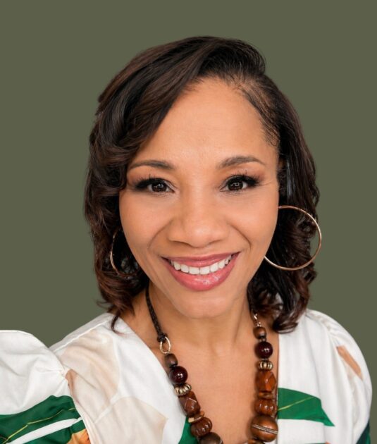Smiling woman with large hoop earrings and beaded necklace, wearing a white patterned blouse, against an olive backdrop.