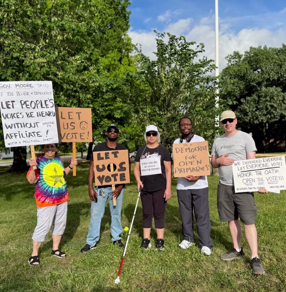 Six protesters stand on grass holding cardboard signs urging voting rights and open primaries, with trees in the background.