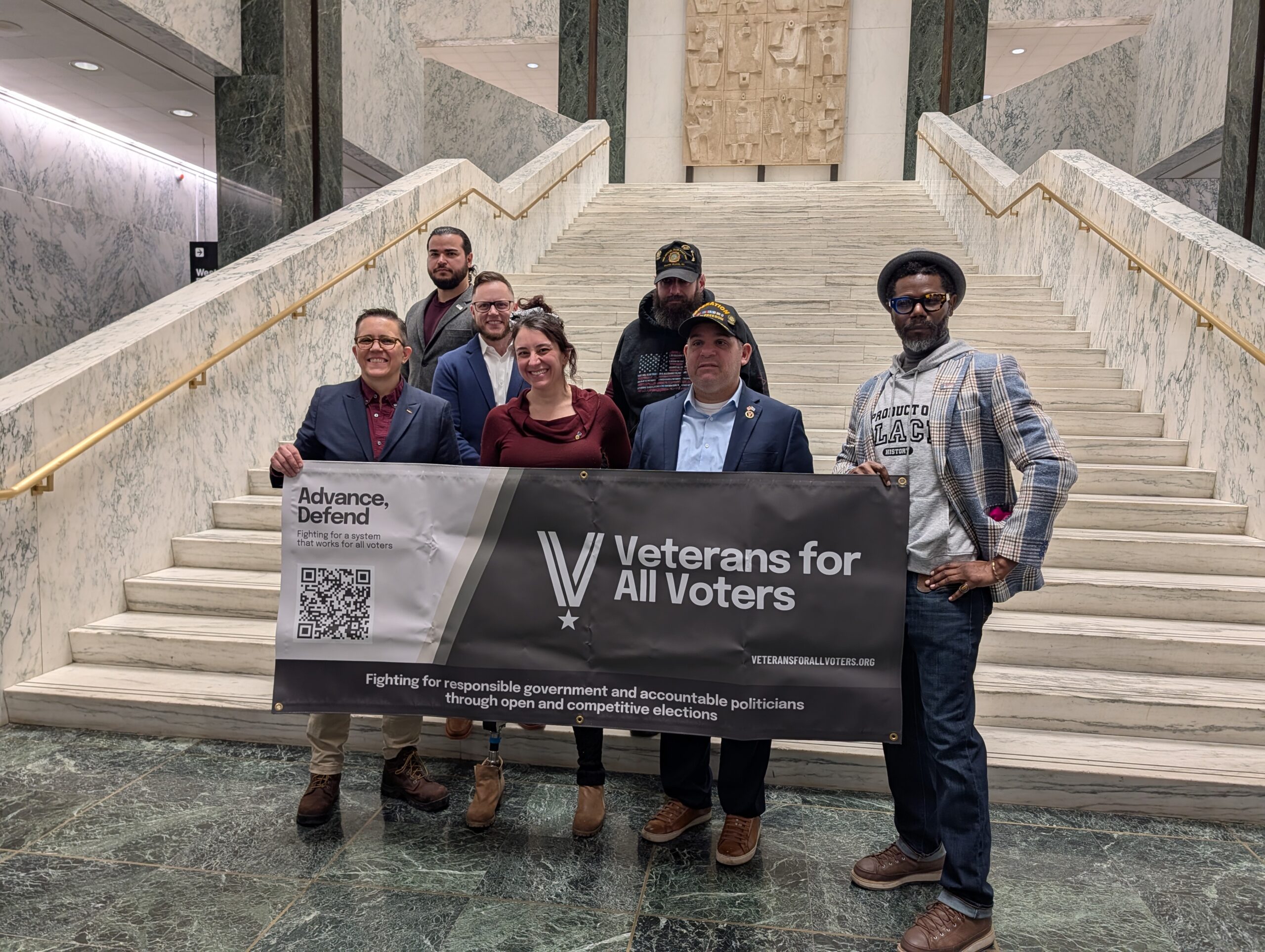 Six people stand on marble stairs behind a banner reading 'Veterans for All Voters' inside a grand building.