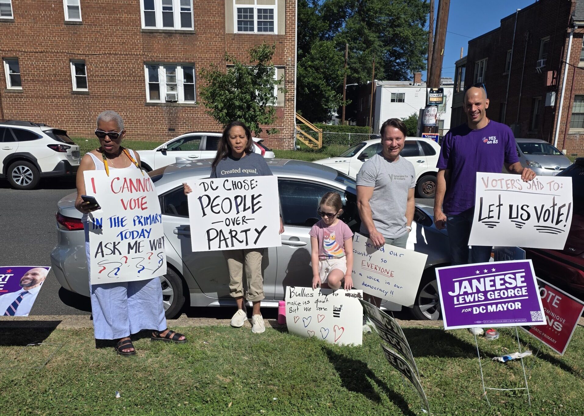 Group of diverse protesters standing on a city street holding signs next to a parked car.
