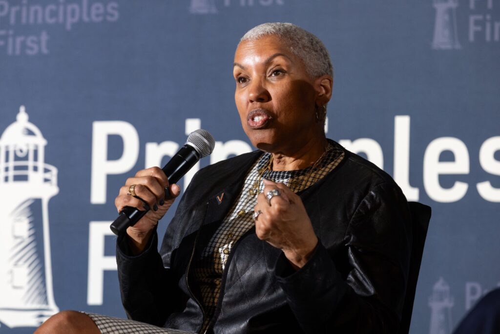 Woman with short gray hair speaking into a handheld microphone on a blue backdrop, panel setting visible in the background.