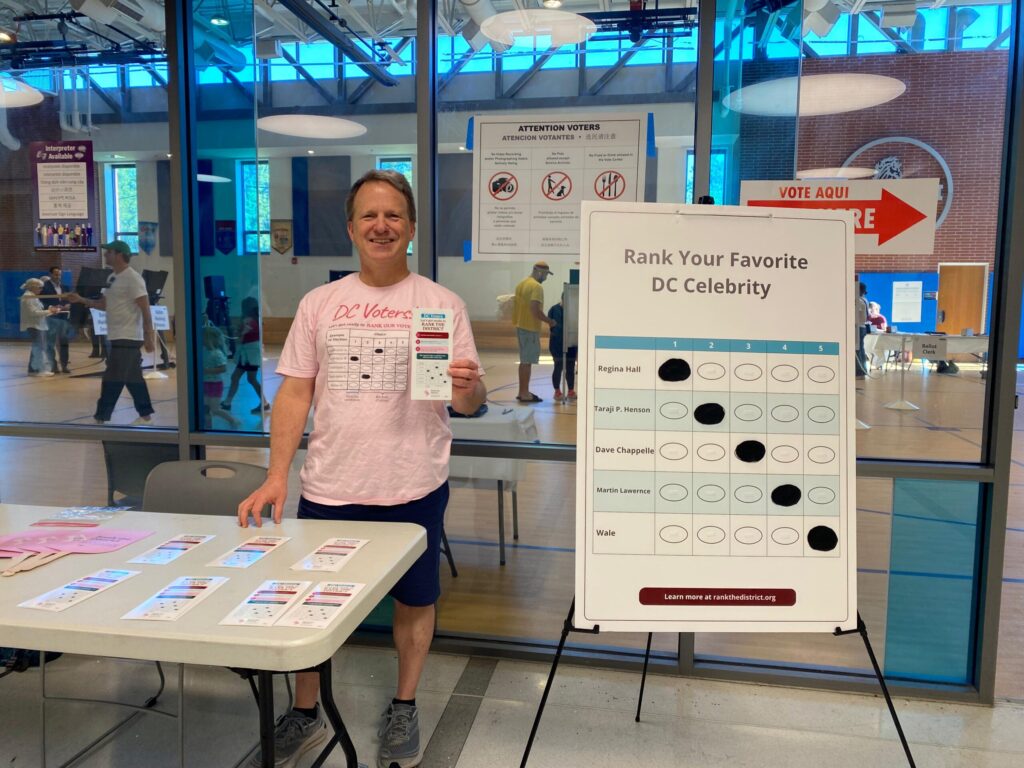 Man in pink T-shirt standing at a table with ballots, signage reading 'Rank Your Favorite DC Celebrity' at a voting/event booth inside a gym-like space.