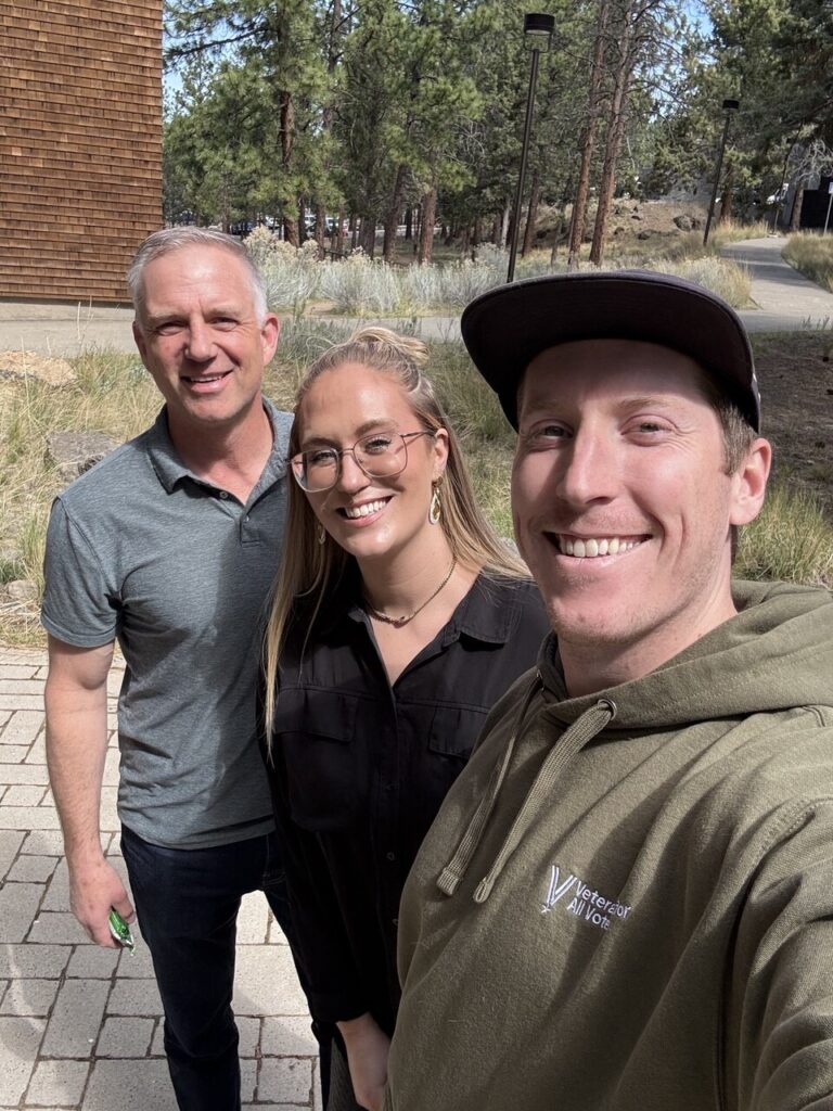Three friends smiling for a selfie outdoors on a sunny day, with trees and a path in the background.